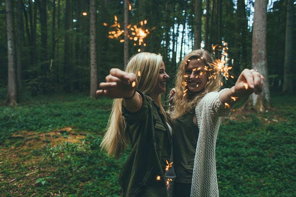 Dos mujeres sosteniendo bengalas se sonríen mientras están de pie en un bosque, capturando un momento alegre de celebración en tu vida.