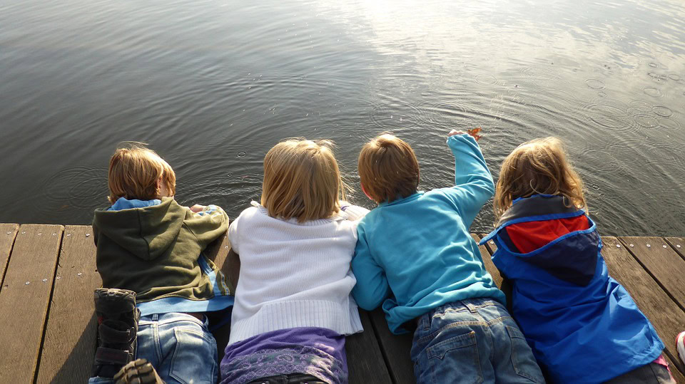 Cuatro niños yacen en un muelle de madera, mirando por encima del borde el agua. Están vestidos con ropa informal y de espaldas a la cámara, un momento sereno que recuerda a los padres la importancia de educar y cuidar mientras encuentran formas de motivar a tus hijos.