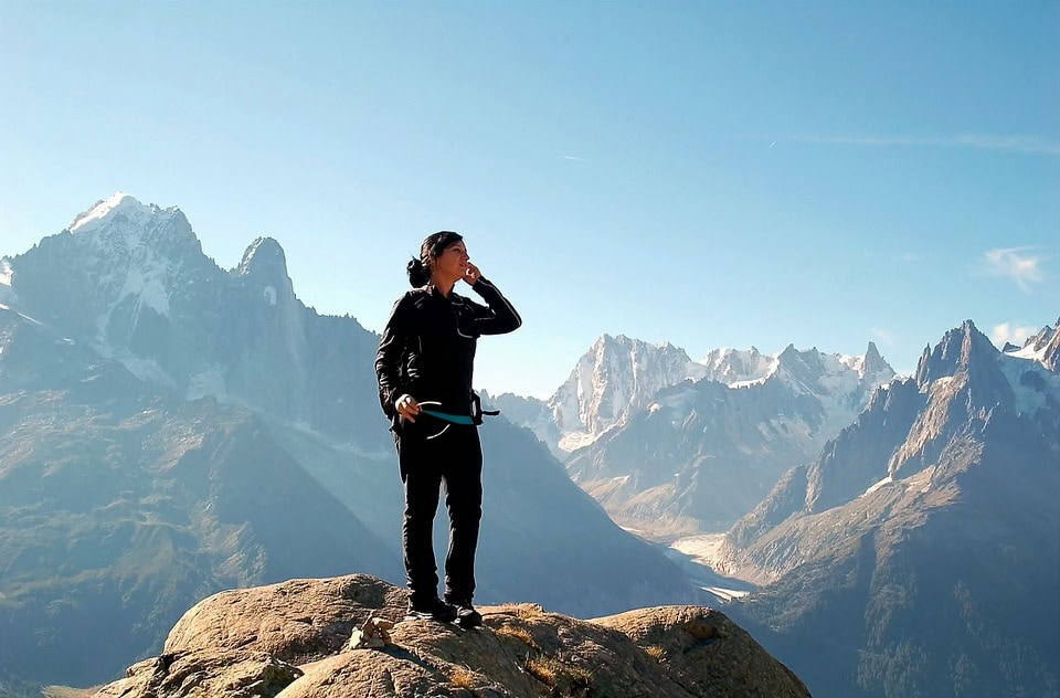 Persona parada en un pico rocoso, sosteniendo un equipo de escalada y contemplando un paisaje montañoso con picos nevados al fondo bajo un cielo azul claro, tal vez reflexionando sobre la elaboración de pensamientos internos o navegando en un duelo personal en medio de la grandeza de la naturaleza.