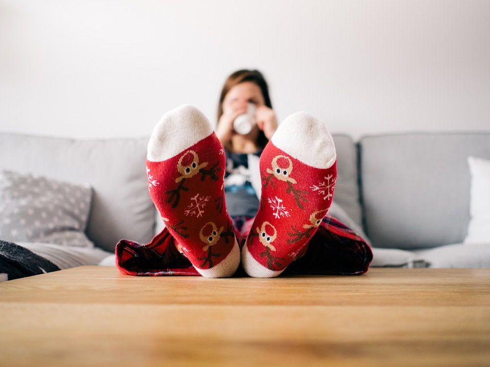 Una persona que lleva calcetines rojos con motivos navideños y diseños de renos está sentada en un sofá gris, con los pies apoyados en una mesa de madera, bebiendo de una taza blanca.