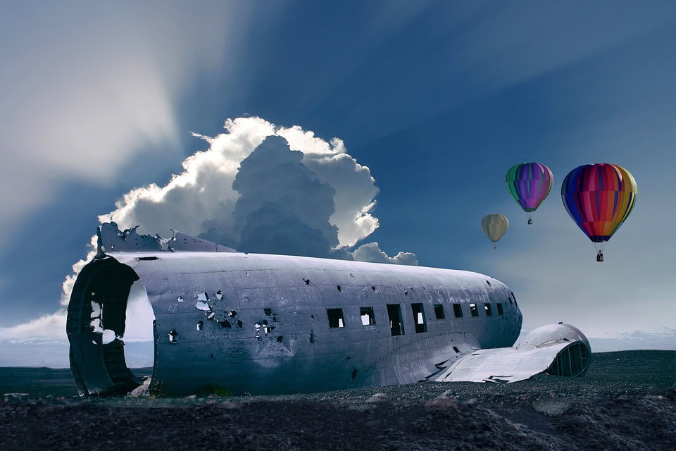 Un fuselaje de avión abandonado y en ruinas se encuentra en un paisaje árido bajo un cielo parcialmente nublado. Descubra tres coloridos globos aerostáticos flotando en el fondo, presentando un marcado contraste con la desolada escena.