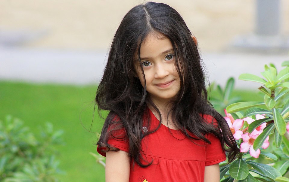 Una joven con cabello largo y oscuro y una camisa roja se encuentra afuera, cerca de plantas verdes y flores rosadas, mostrando la fórmula perfecta entre la belleza y la inocencia de la naturaleza.