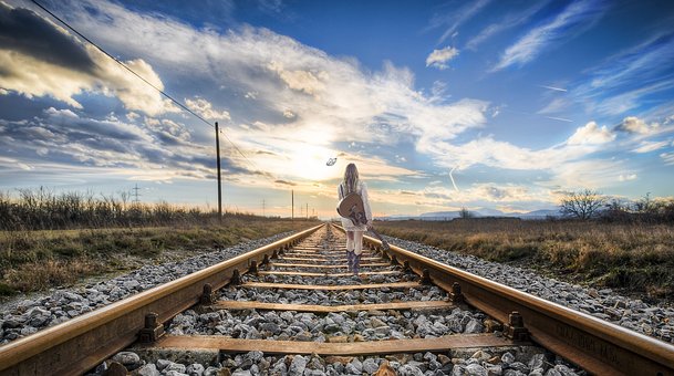 Una persona con cabello largo camina a lo largo de una vía de tren durante el atardecer, decidiendo su camino mientras sostiene una guitarra acústica, con campos y líneas eléctricas a ambos lados.