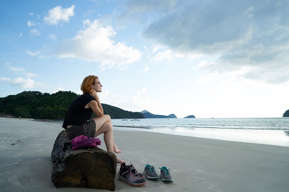 Una persona sentada en una roca junto a la playa, mirando el océano con zapatos colocados en la arena cercana. El cielo está parcialmente nublado y las islas lejanas se ven al fondo, ofreciendo un entorno sereno para la contemplación en tiempos de duelo.