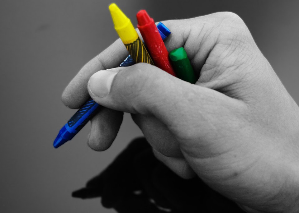 Una mano que sostiene cuatro crayones en azul, amarillo, rojo y verde se destaca sobre un fondo blanco y negro, resaltando lo que no sabes sobre tus necesidades en la expresión creativa.