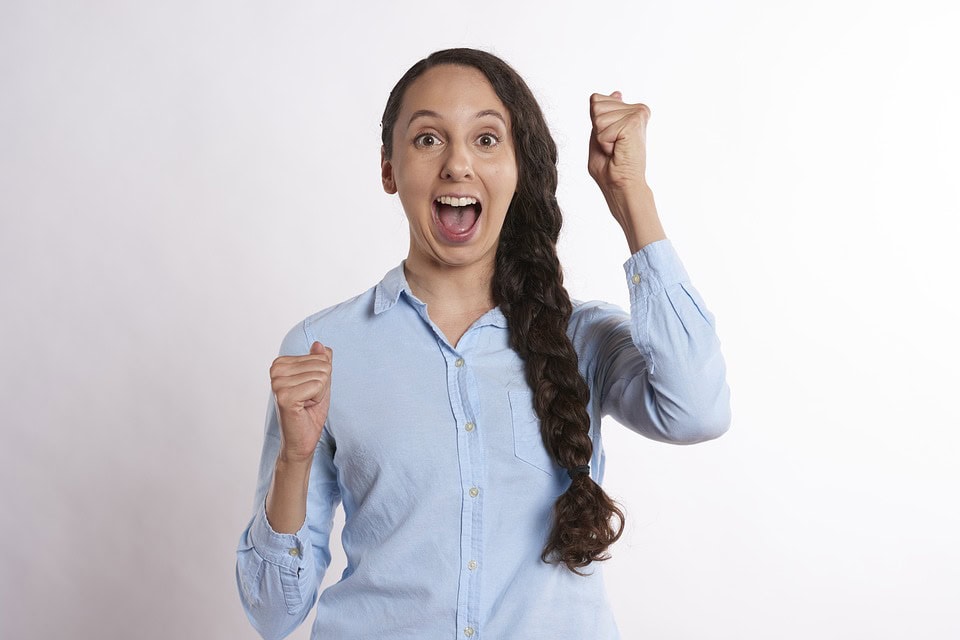 Una mujer con el pelo largo recogido en una trenza, vestida con una camisa azul, aparece emocionada con la boca abierta y los puños cerrados, celebrando como si estuviera lista para atrapar el éxito sobre un fondo liso y blanco.