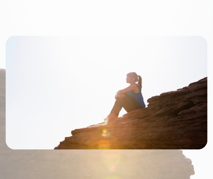 Persona sentada sobre un saliente de roca en posición sentada, aparentemente perdida en sobrepensar, con el sol brillando intensamente de fondo.
