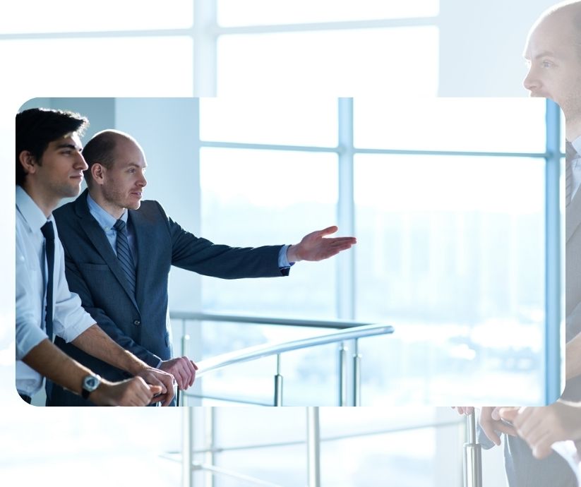 Tres hombres vestidos de negocios se encuentran en una oficina bien iluminada con ventanales del suelo al techo. Un hombre hace un gesto con la mano, sugiriendo que están conversando sobre estrategias para reducir la ansiedad y promover la seguridad psicológica en el trabajo.