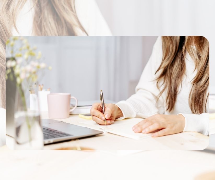 Una persona de cabello largo escribe afirmaciones positivas en un cuaderno con un bolígrafo, sentada en un escritorio con una computadora portátil, una taza rosa y un jarrón de flores. Afirmaciones para Tener Confianza en Sí Mismo
