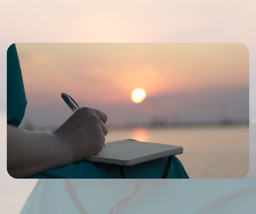 Persona escribiendo en un cuaderno al atardecer con un fondo borroso de un cuerpo de agua y cielo, encontrando consuelo en la vuelta a la rutina después del verano.