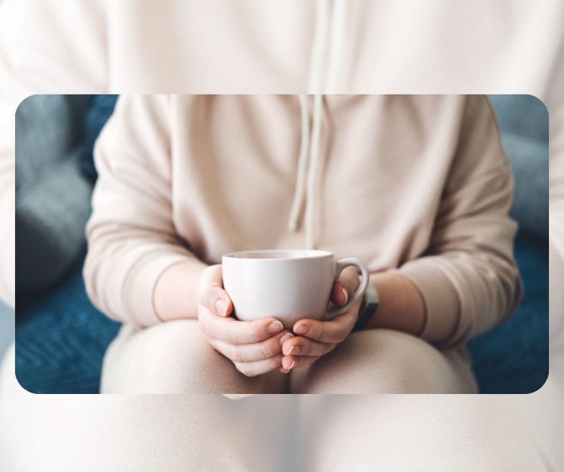 Una persona que viste una sudadera de color beige sostiene una taza de cerámica blanca con ambas manos mientras está sentada, encontrando consuelo en una de sus herramientas para reducir el estrés.