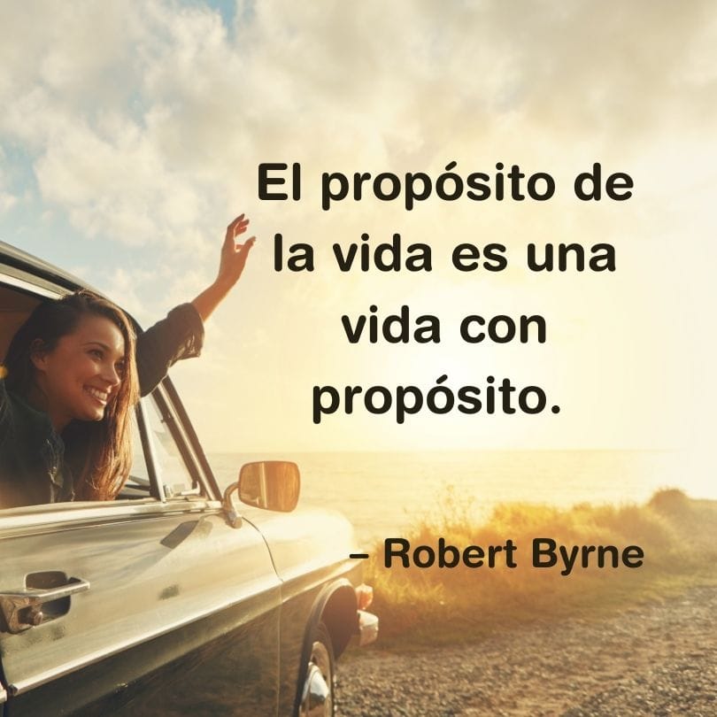 Una mujer saluda desde la ventanilla de un coche en la playa. La cita en español de Robert Byrne dice: "El propósito en la vida es una vida con propósito".